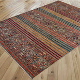 A rectangular Afghan Loribaft rug with a traditional geometric pattern and plain modern stripes in red, green, gold, and orange tones, shown laid out on a light wood plank floor.