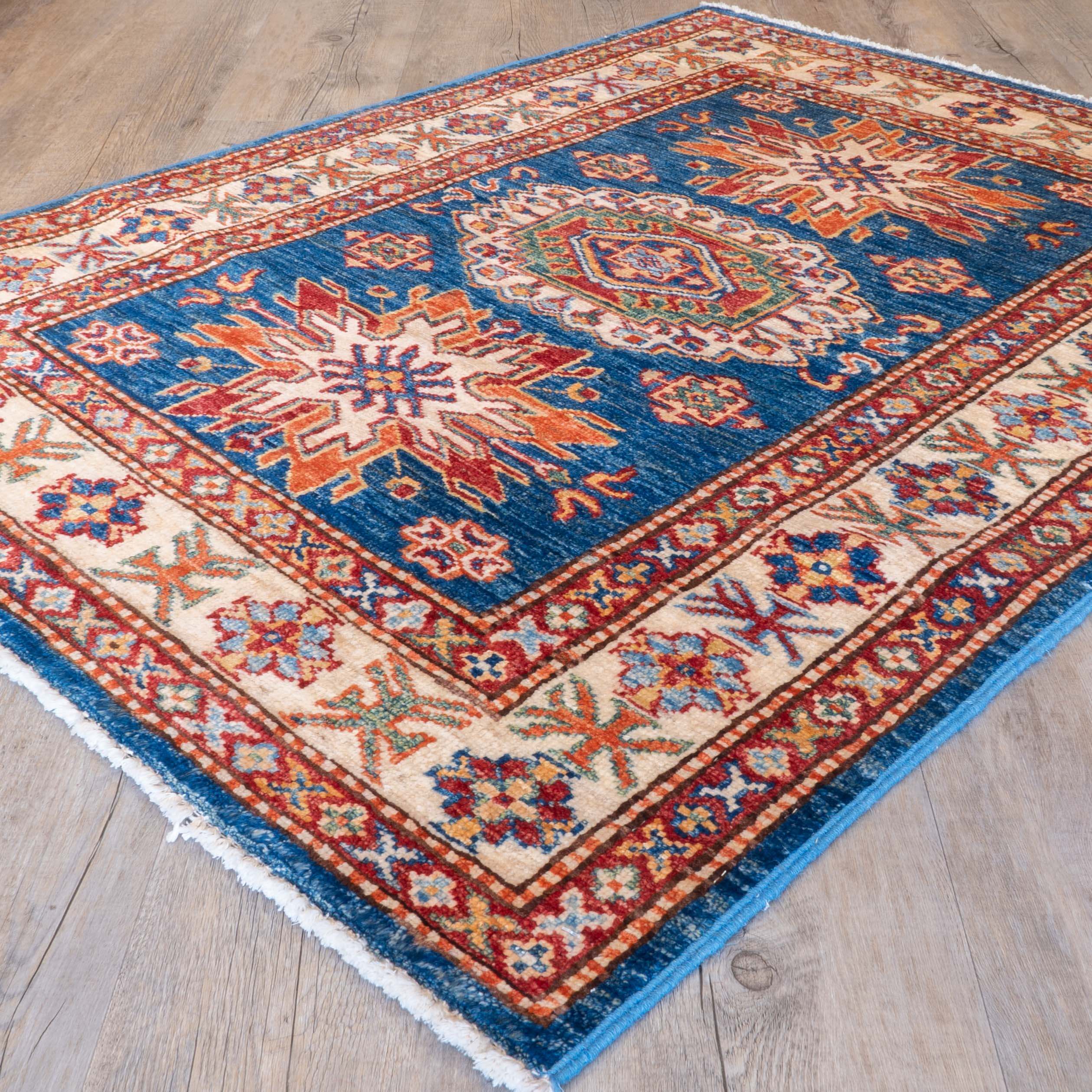 A traditional geometric patterned rug with a blue central field, cream borders, and red, orange, and green accents, shown laid out on a light wood floor.