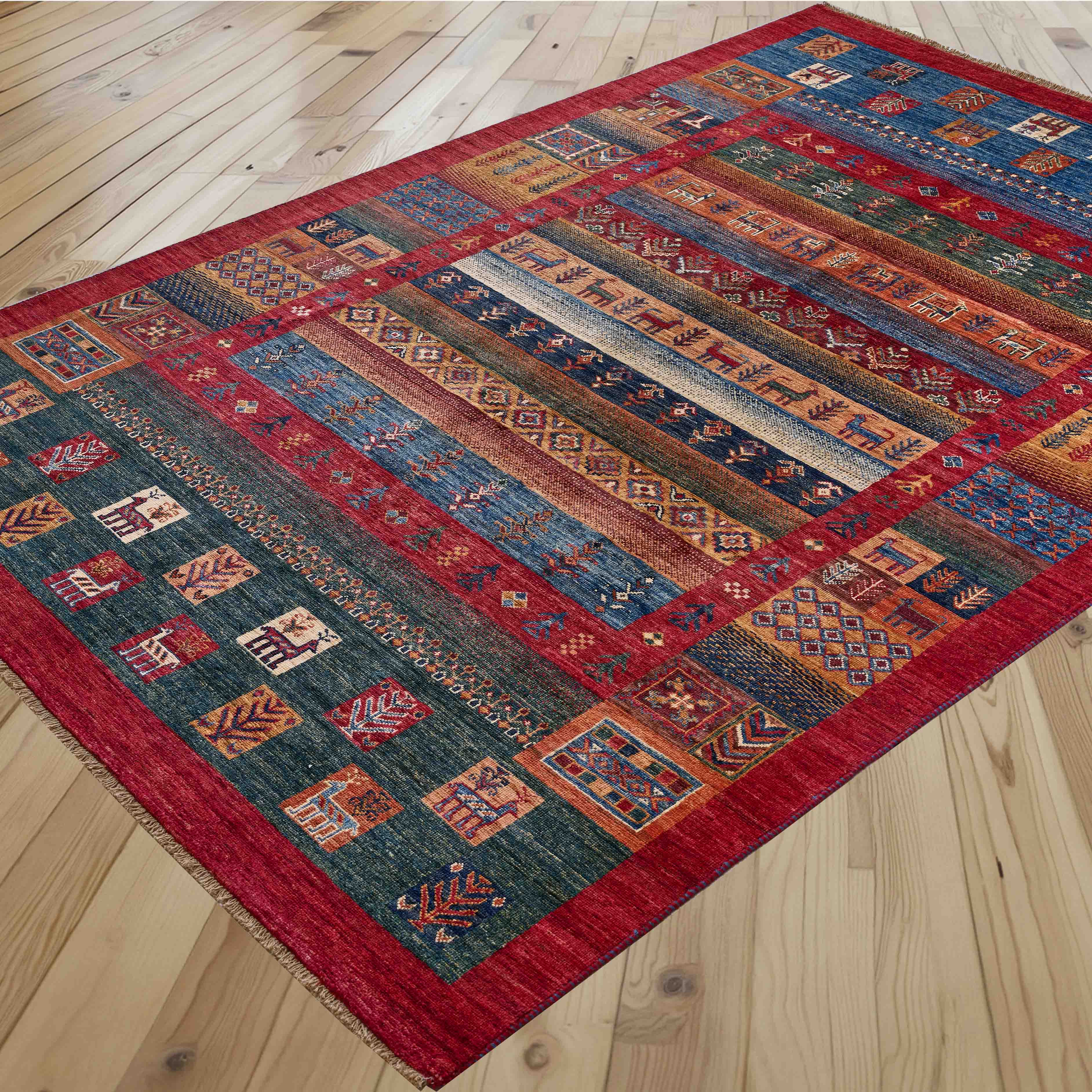 A rectangular area rug with a complex geometric and tribal pattern in red, blue, green, and beige, laid out diagonally on a light-colored wooden floor.