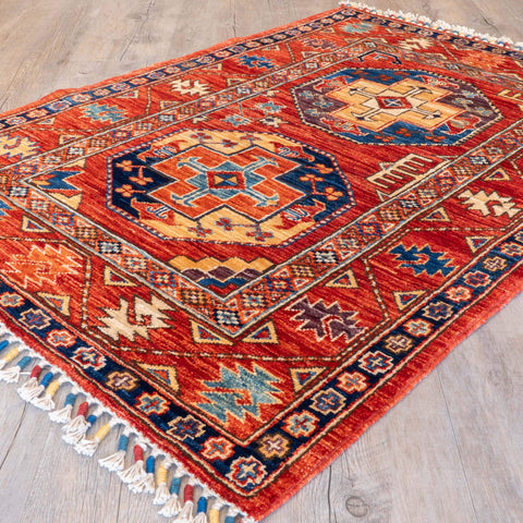A traditional hand-woven Afghan rug with a vibrant red-orange base, featuring two large central medallions in navy blue, gold, and orange, and geometric borders. The rug is shown laid out on a light-colored wooden floor, with white fringed tassels visible on the left and bottom edges.