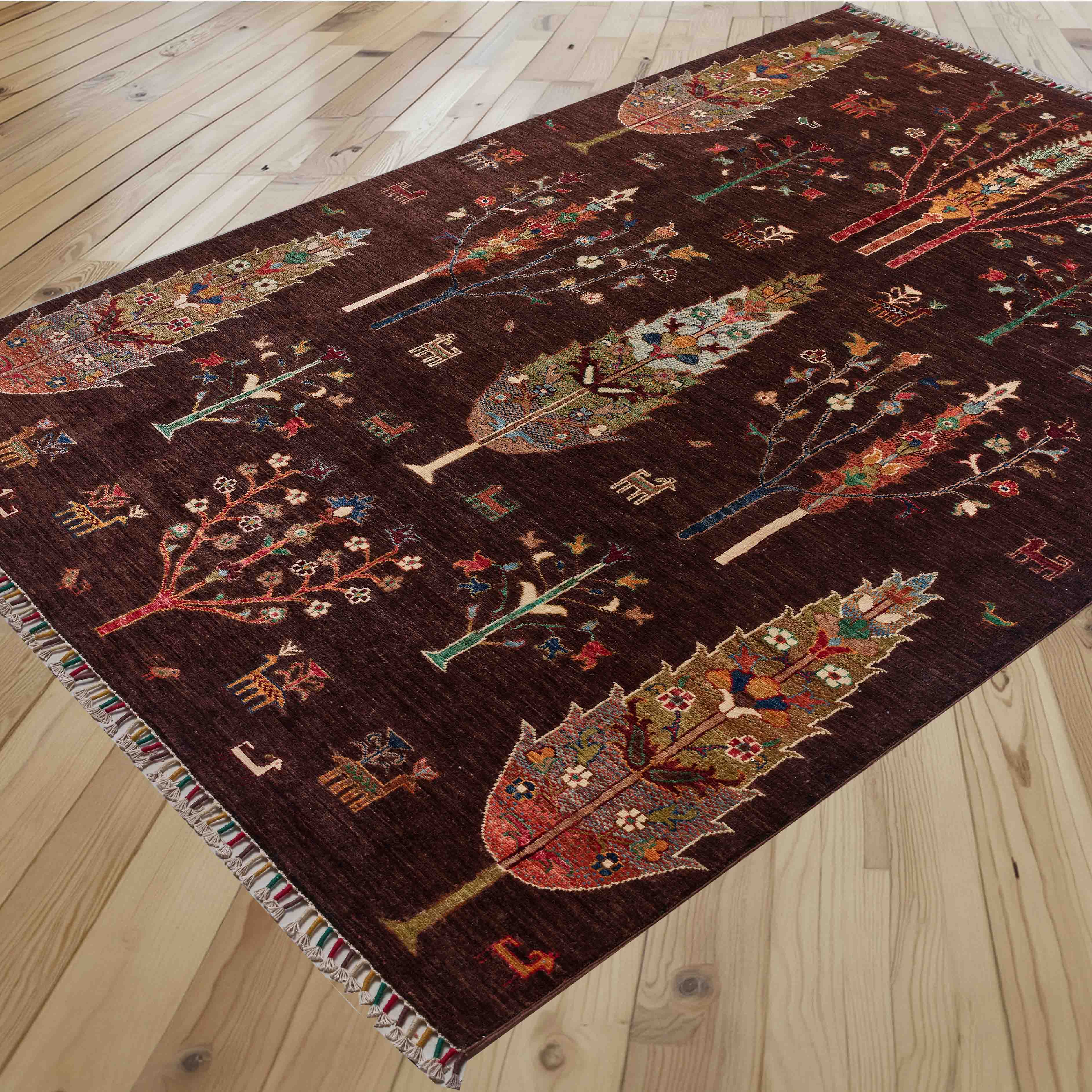 A dark brown area rug with a detailed, colourful tribal tree pattern sprinkled with stylised animals and birds, laid out on a light-colored wooden floor.