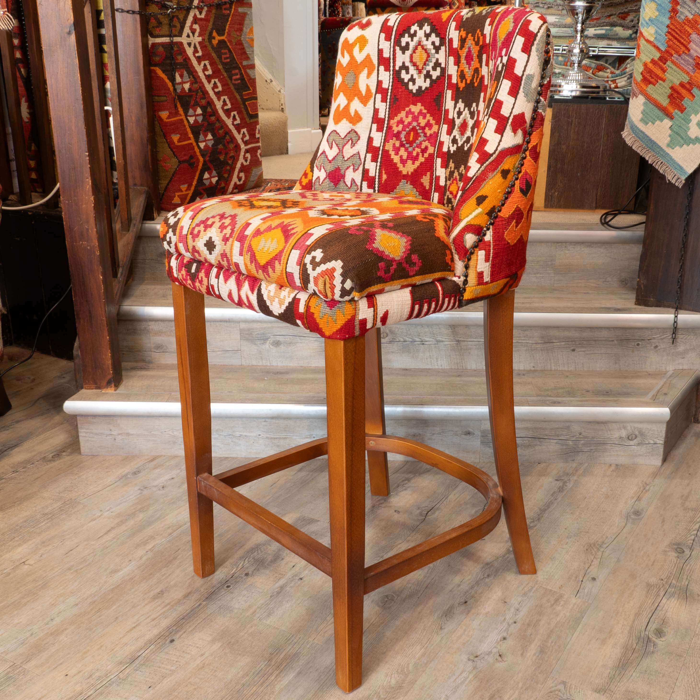 A wooden bar stool with a colorful geometric patterned fabric seat and backrest, set on a light wood floor in front of indoor stairs at Jenningsrugs