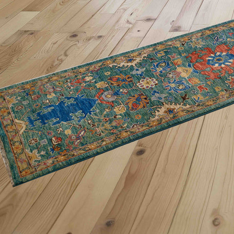 A long runner rug with a distressed traditional pattern in teal, blue, papaya, and honey, laid out diagonally on a light-colored wooden plank floor.