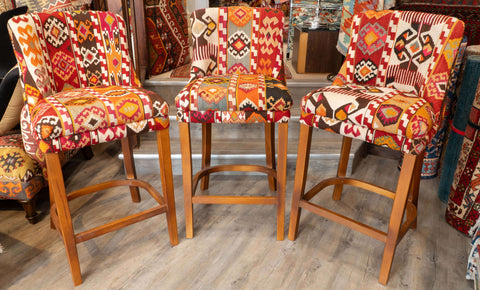 Three wooden bar stools with colourful geometric patterned upholstery in red, orange, beige, and black, displayed on a wood-look floor in a Jennings rug store.