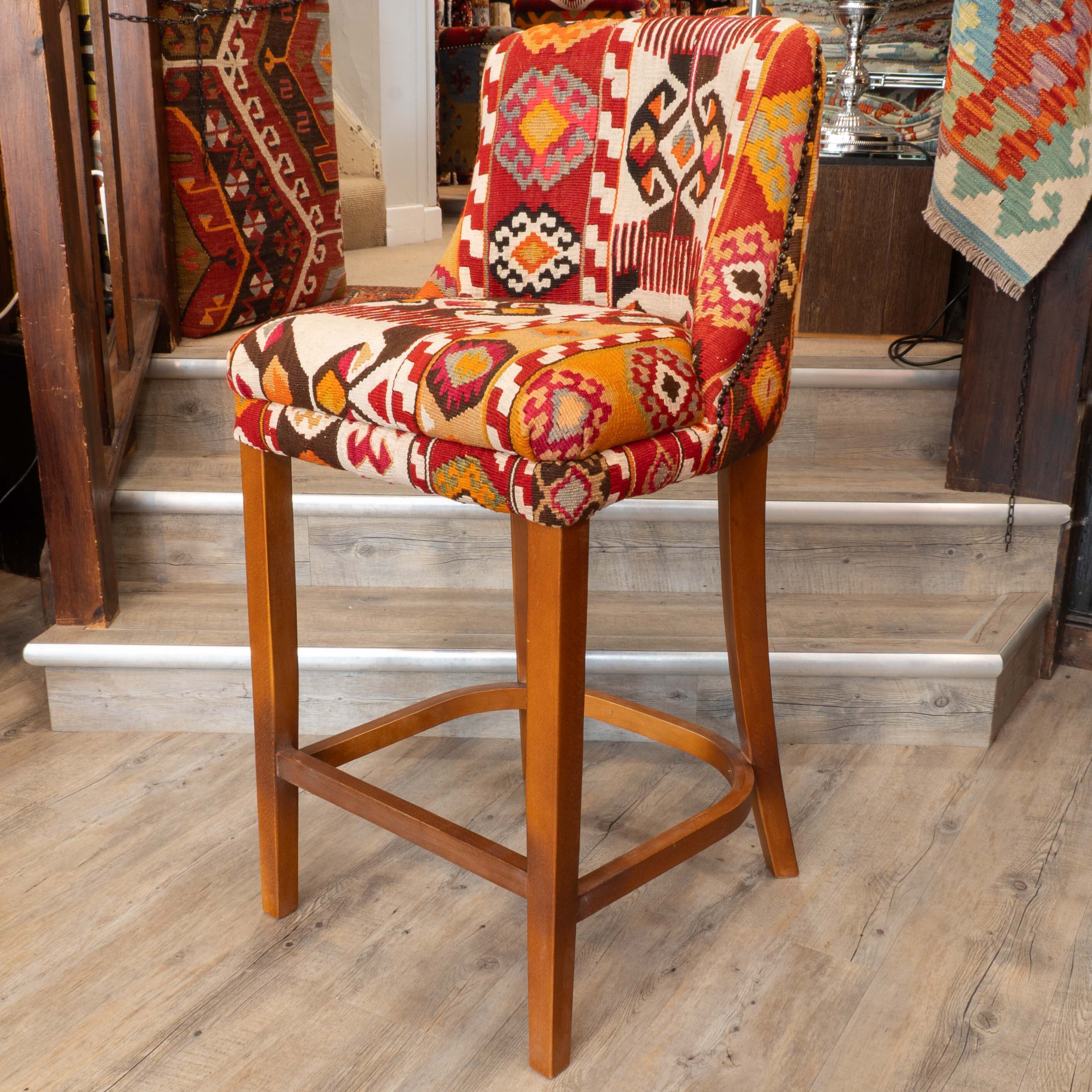 A wooden bar stool with a colorful geometric kilim patterned seat and backrest, set in a home interior with wooden stairs and rugs in the background.