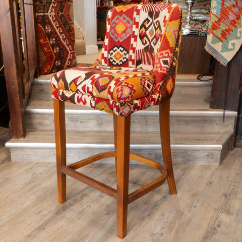 A wooden bar stool with a colorful geometric kilim patterned seat and backrest, set in a home interior with wooden stairs and rugs in the background.