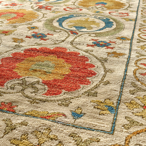 Close-up of a woven area rug with an ornate floral and scroll pattern in red, yellow, blue, and olive on a warm cream background.