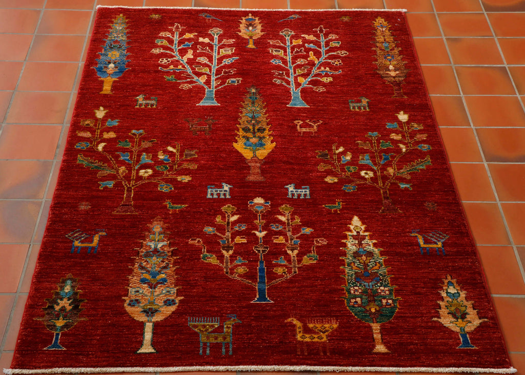 This is a hand knotted rug woven in Afghanistan with a wool pile. The foundation is cotton and it has a short cotton fringe. The background colour is a beautiful rich red and the design is a fairly primitive one with differing styles of trees and random stylised goats and birds. The other colours are old gold, sand, tan, and different shades of blue including turquoise .