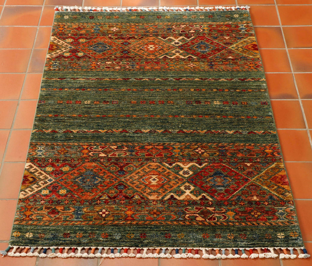 This is a small rug sized 121 by 77 centimetres.Wonderful colours used in this Afghan Samarkand rug with the main colour being a mottled racing green shade coupled with rust, gold, peach, cream and blue tones in the traditional design on the ends of the rug.   There are two large bands of decoration at either end of the rug incorporating various stylised flowers, hooks and large rhombi with internal decoration..  This piece also has the coloured wound fringes.