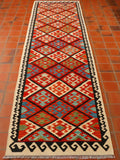 This striking Afghan Kilim runner is a mix of wonderful colours that really bring it to life and make it sing. The outer border is oatmeal colour with a deep navy pattern which really stands out and frames the runner. The inner planner is covered with large diamonds in colours of flame orange, mid blue, olive green and turquoise. In this photograph the runner is sitting on terracotta tiles and looks really great.